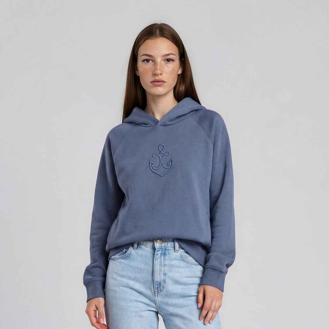 A woman stands barefoot against a light gray studio background, wearing a dusty blue cropped hoodie with a tonal embroidered logo on the chest and light-wash blue jeans.