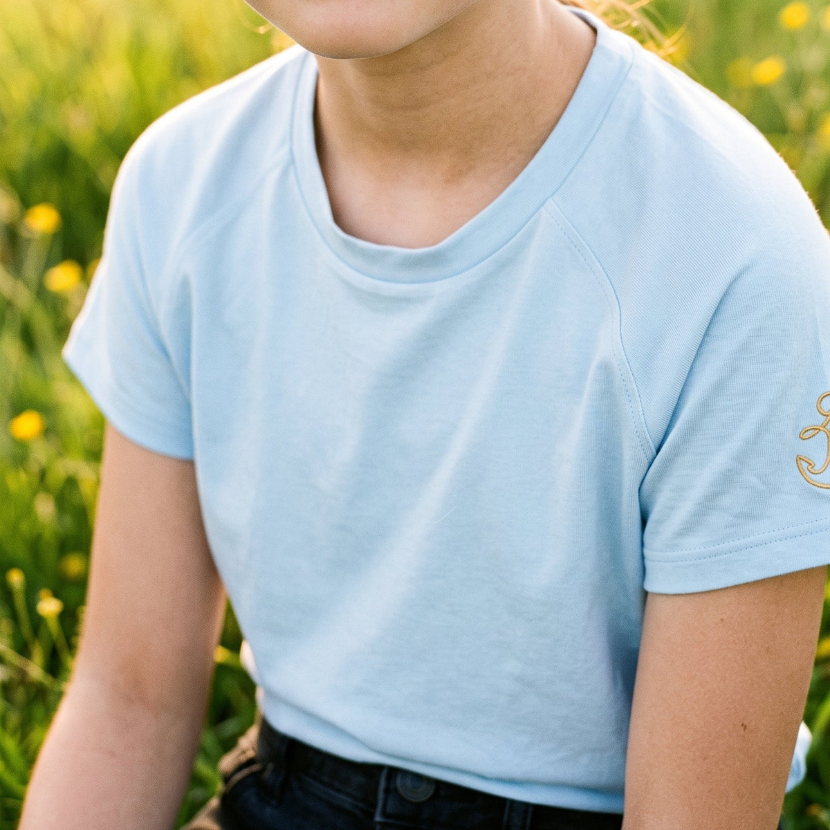 A young girl with brown hair tied back, wearing a light blue short-sleeve t-shirt and dark jeans, sitting in a grassy field with yellow flowers.